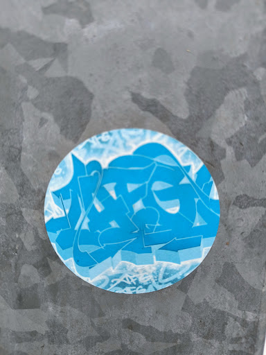 A round sticker with light blue and blue graffiti art. The sticker is affixed to a gray metallic surface. No visible text or QR code.