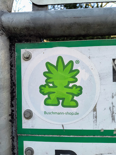 A round sticker featuring a stylized green character with cannabis leaf-shaped hair.  The text "Buschmann-shop.de" is visible below the character.