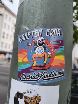 Street sticker The sticker features a cartoon character resembling a rabbit with large, round eyes, one with a blue iris and the other a black pupil, and a wide, toothy grin. It has long ears and is wearing a purple beanie. The character is depicted in a dynamic pose, riding a red skateboard. It's holding a small lantern with a glowing yellow light in its right hand. The background is a vibrant blue with a rainbow-colored arc behind the character, radiating outwards with jagged lines and stars. Below the character, the text 'Electric Kindieland' is written in a stylized, bold font with a black outline. At the very bottom, a website address is visible.