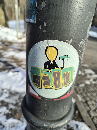 Street sticker A round sticker is affixed to a dark gray pole. The sticker has a light blue background and a white border. Centered on the sticker is a black figure with a yellow smiley face. The figure holds a paint roller in its hand, with blue paint dripping from the roller. Below the figure, the word "SEDQ" is written in green and yellow block letters.