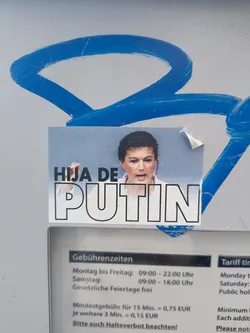 A rectangular sticker adhered to a metallic surface features a photo of a woman and the text "HIJA DE PUTIN" in bold, outlined letters. The woman in the photo is giving a fist salute. The background is slightly blurred and indistinct, while a portion of a sign with German text is visible beneath the sticker.