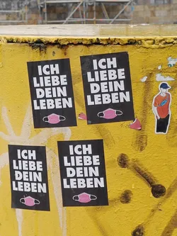 Street sticker Four identical stickers are affixed to a yellow wall. Each sticker has black background with white text that reads 'ICH LIEBE DEIN LEBEN', which translates from German to 'I love your life' in English. Each sticker also contains a small pink face mask graphic at the bottom. The stickers are slightly worn, suggesting they have been exposed to the elements.