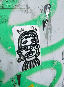 Street sticker The sticker features a quirky black and white drawing of a character with four eyes, large lips, and a striped shirt. The text "Rodo Dodo" is written above the head of the character. It is pasted on a rough surface with green graffiti.