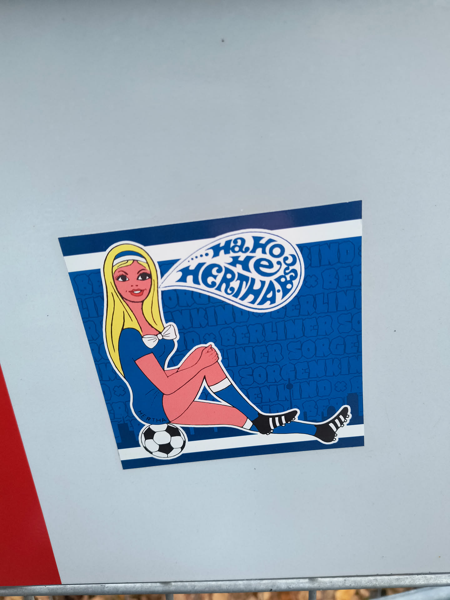 The sticker is predominantly blue and white, with a central illustration of a blonde woman with large eyes and red lips, wearing a blue soccer jersey with a white bow tie, pink shorts, and striped socks. She is sitting with her legs crossed, one foot resting on a black and white soccer ball. A speech bubble above her head contains the text 'Ha ho HERTHA!' in a stylized blue font. The background of the sticker is a dark blue with a repeating pattern of the word 'BERLINER' and other illegible text, interspersed with small star-like symbols. The overall style is reminiscent of vintage comic or pop art.