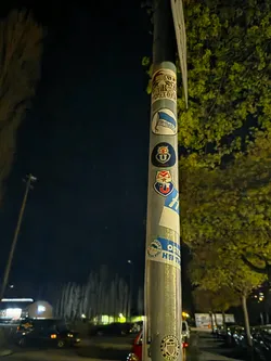 Street sticker The image centers on a metal pole covered with a vertical arrangement of multiple circular and oval stickers. The stickers are layered and show signs of wear, suggesting they are genuine physical decals. From top to bottom, visible stickers include a partially obscured design featuring a skull-like figure with red, white, and black elements, and text 'OSTBER'. Below this is an oval white sticker with a blue flag-like shape and 'Hertha BSC' written in blue. Following this are two circular stickers, one with a dark blue background and the other red, both depicting an owl-like emblem and the word 'UNION', likely representing 1. FC Union Berlin. Further down, a blue and white sticker shows partial text 'OST HEI TH'. At the bottom, a circular white sticker with black vertical stripes and a crest is visible, encircled by the text 'S.V. NORMANNIA BERLIN 08'. The overall appearance is a collection of fan or club emblems, predominantly in blue, white, red, and black colors, against the metallic grey of the pole.