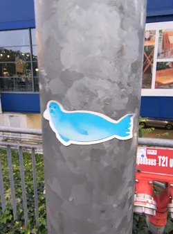 Street sticker The sticker features a stylized, cartoonish seal in a vibrant shade of blue. It is depicted in profile, facing left, with its body elongated and streamlined. The seal has a small, rounded head with subtle facial features, including two small eyes and a hint of a mouth. Its flippers are suggested by simple lines. The tail is fanned out with distinct, rounded lobes. The entire image is outlined with a thick white border, giving it a distinct sticker appearance.