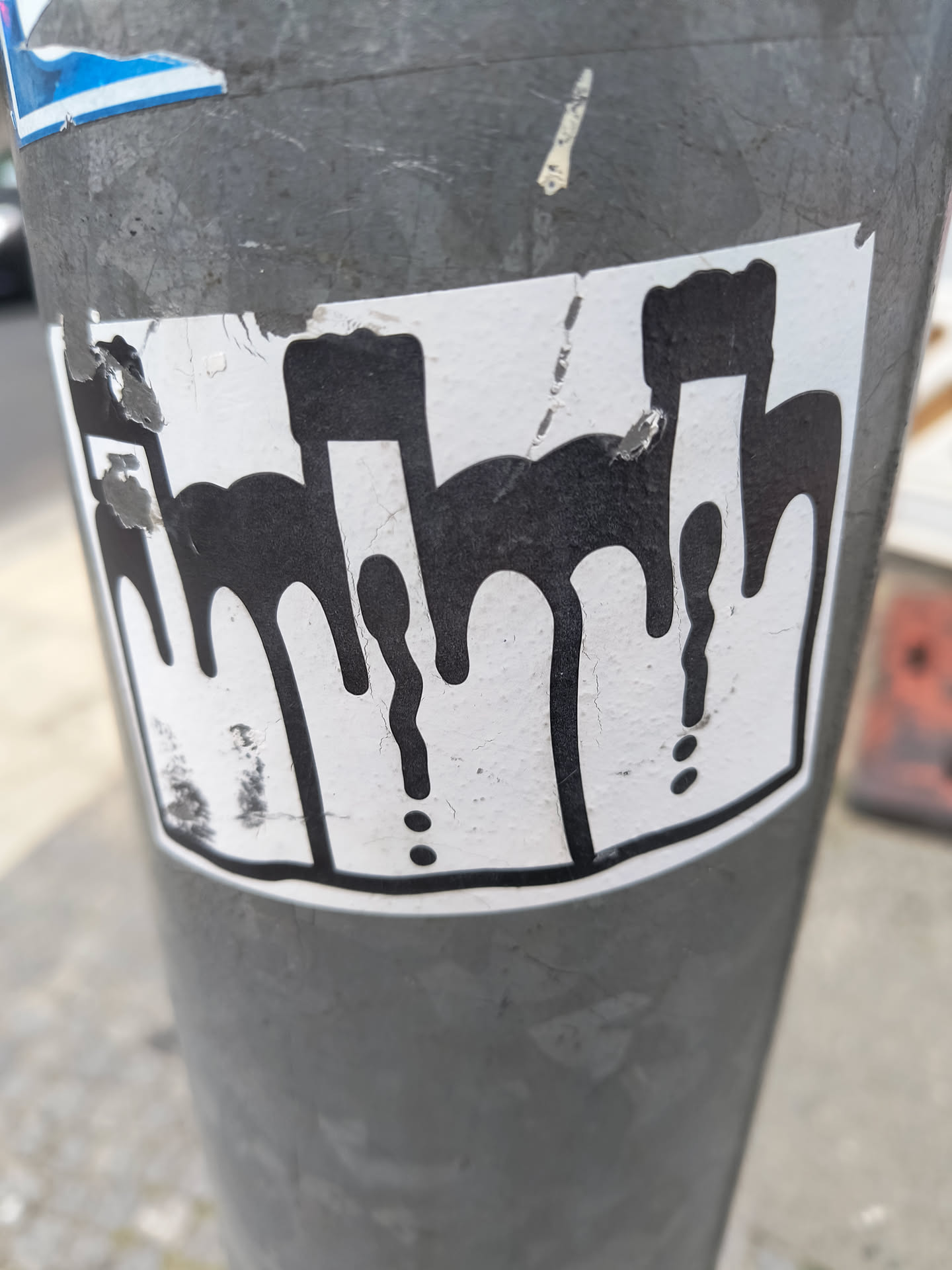 A sticker with a black outline and dripping shapes. The shapes are arranged horizontally and the dripping effect is pronounced. The background is white and the sticker appears to be affixed to a metal pole.