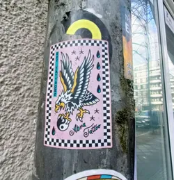 Street sticker A sticker featuring a traditional American eagle tattoo design holding a yin yang symbol. The sticker has a pink background with a checkered border and text that says "Abra Tattoo" and "GET TATTOOED".