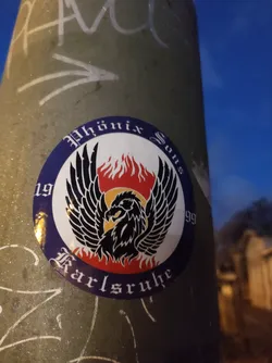 Street sticker Round sticker featuring a black phoenix with outstretched wings, set against a backdrop of flames. The text 'Phönix Sous' is written in a stylized font above the phoenix, while '1999' is visible on the left and 'Karlsruhe' at the bottom, also in stylized fonts. The colors are primarily blue, red, yellow, and black.