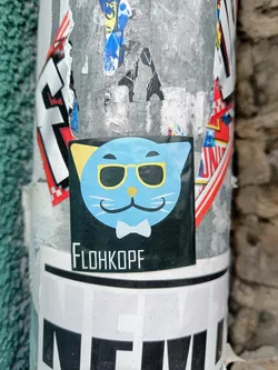 Street sticker A black sticker with a cartoon illustration of a light blue cat wearing sunglasses and a bow tie. The text 'FLOHKOPF' is visible at the bottom of the sticker. The sticker is affixed to a pole covered in other stickers and graffiti.