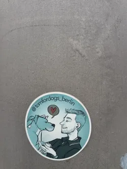 Street sticker The sticker is circular with a white border. It features a stylized illustration of a man and a dog. The man is on the right, depicted in profile with his eyes closed and a smile, wearing a dark collared shirt. The dog, on the left, is a light blue pitbull-type breed, also in profile, with its eyes closed and a red heart symbol in a speech bubble above its head. The background is a gradient of light blue. The text '@tomfordogs_berlin' is written in a curve along the top edge of the sticker.