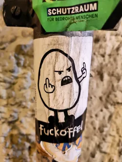 A sticker featuring a simple drawing of an angry potato-like character flipping off the viewer. The sticker has the text "Fuckoff" written at the bottom.  It is stuck to a pole, partially obscuring other stickers underneath.