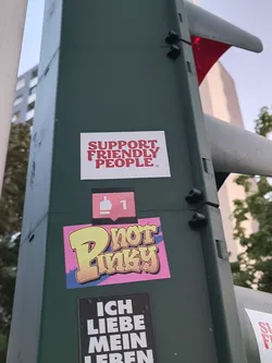 Street sticker The sticker is composed of three distinct elements layered on a dark green pole. The topmost element is a white rectangular sticker with bold red text that reads 'SUPPORT FRIENDLY PEOPLE.' Below this, a smaller sticker displays a pink speech bubble icon with a white thumbs-up symbol and the number '1' inside, resembling a social media 'like' notification. The largest and most vibrant sticker is situated at the bottom, featuring the word 'Pinky' in a graffiti-style font. The letters are yellow with a textured gradient, outlined in black and white, and set against a bright pink background with blue and purple abstract shapes. Another black sticker with white text is partially visible below the 'Pinky' sticker.