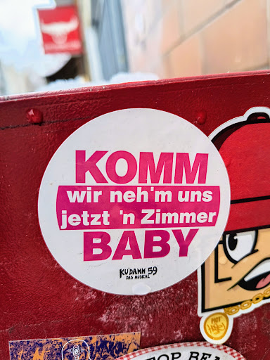 Round white sticker with pink text on a red surface. The text says "Komm wir neh'm uns jetzt 'n Zimmer Baby" which translates from German to "Come let's take a room now baby".  There is small text at the bottom that says "Kudamm 59 das Musical".