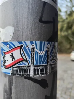 Street sticker The sticker features a stylized depiction of the Olympic Stadium in Berlin, with the Olympic rings prominently displayed above it. To the left, the flag of Berlin, featuring a black bear on a white and red background, is shown waving. The background of the sticker consists of radiating blue and white stripes, reminiscent of a sunburst or stadium lights. Several soccer balls are scattered across the top of the sticker, reinforcing the football theme. The overall design is bold and graphic, with clear outlines and contrasting colors.