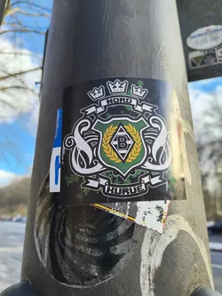 This is a black sticker on a metallic post featuring the Borussia Mönchengladbach logo. The logo has a black background with a green and white shield in the center containing a black 'B' on a white diamond. The shield is surrounded by a gold laurel wreath and flanked by two white swans. Above the shield are three crowns, and banners above and below read 'NORD' and 'KURVE' respectively.