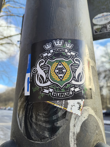 Street sticker This is a black sticker on a metallic post featuring the Borussia Mönchengladbach logo. The logo has a black background with a green and white shield in the center containing a black 'B' on a white diamond. The shield is surrounded by a gold laurel wreath and flanked by two white swans. Above the shield are three crowns, and banners above and below read 'NORD' and 'KURVE' respectively.