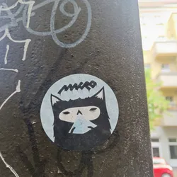 A round sticker featuring a stylized black cat with the word "MKRO" above it. The cat has a mask-like face and appears to be wearing a mask or hood. The background of the sticker is light blue. The sticker is affixed to a dark-colored surface that appears to be outdoors.