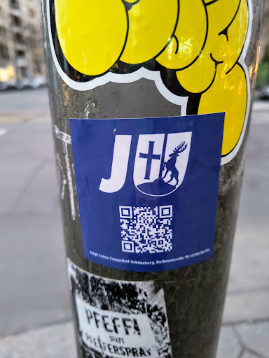 Street sticker A blue square sticker with a white logo. The logo contains a stylized 'J+' with a cross and a deer emblem underneath.  A QR code is located below the logo. Small text at the very bottom appears to give an address. 