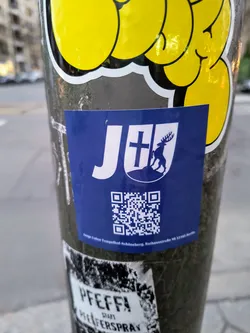 Street sticker A blue square sticker with a white logo. The logo contains a stylized 'J+' with a cross and a deer emblem underneath.  A QR code is located below the logo. Small text at the very bottom appears to give an address. 