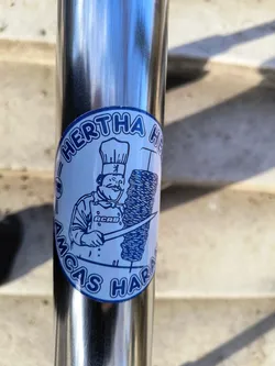 A round sticker with a illustration of a chef holding a knife next to a vertical kebab. The text around says Hertha Herthas Amcas Haras