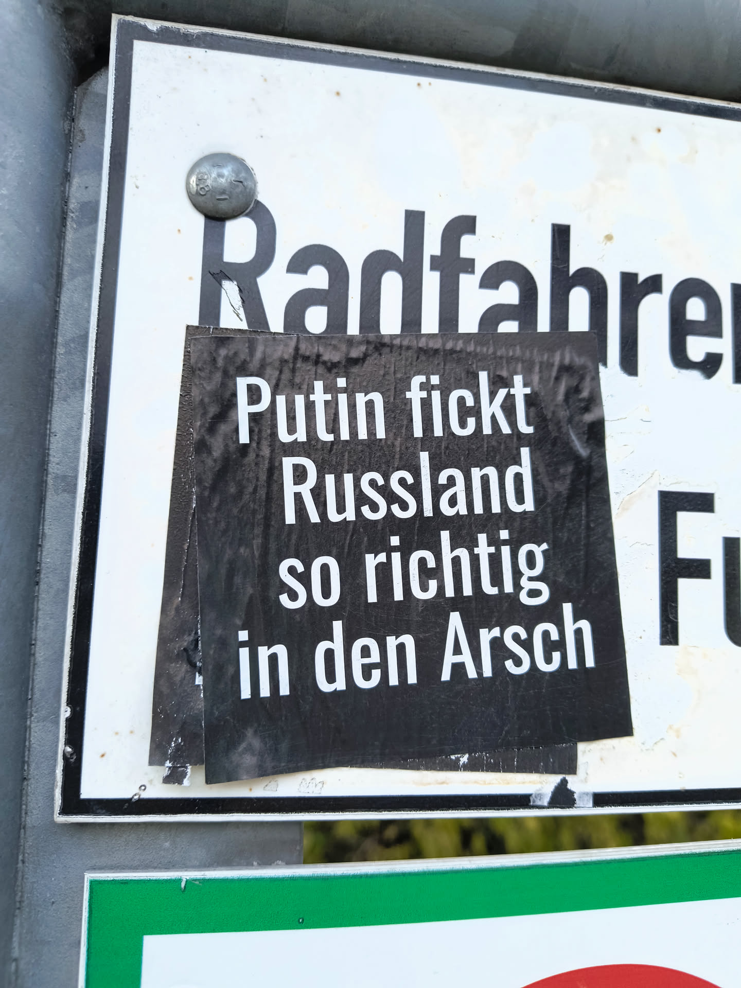 The sticker is black with white, sans-serif text. It is placed centrally on a white sign with black border, which itself is mounted on a metal pole. The sticker appears to be slightly crumpled and has some wear around the edges. The text is in German and is quite explicit.