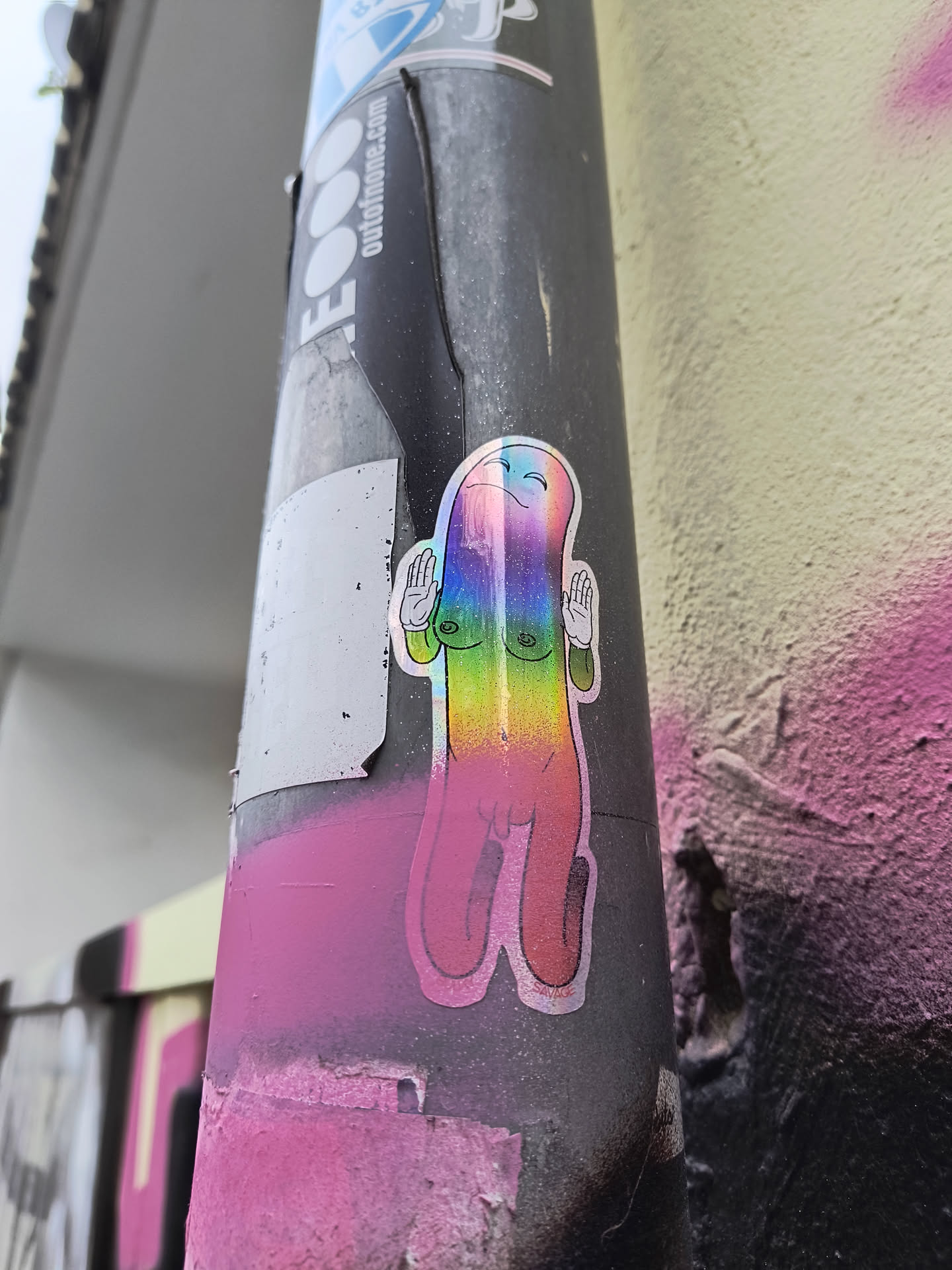 The sticker features a stylized, elongated figure with a rounded top and two distinct legs. The figure is rendered in a holographic material, giving it a shimmering, rainbow-like appearance that shifts with light. It has a simple, smiling face with closed eyes and a blush on its cheeks. Two hands are raised, palms facing outward, in a gesture of greeting or peace. The figure has two small, circular breasts depicted. The outline of the sticker is a thin, dark line. Below the figure, there's a hint of pink spray paint on the surface it's attached to.