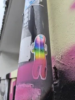Street sticker The sticker features a stylized, elongated figure with a rounded top and two distinct legs. The figure is rendered in a holographic material, giving it a shimmering, rainbow-like appearance that shifts with light. It has a simple, smiling face with closed eyes and a blush on its cheeks. Two hands are raised, palms facing outward, in a gesture of greeting or peace. The figure has two small, circular breasts depicted. The outline of the sticker is a thin, dark line. Below the figure, there's a hint of pink spray paint on the surface it's attached to.