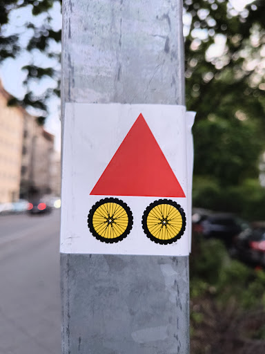 This sticker features a red triangle sitting atop two bicycle wheels. The triangle is equilateral and solid red. The wheels are yellow with black tires and detailed spokes. It's pasted on a grey metal pole.