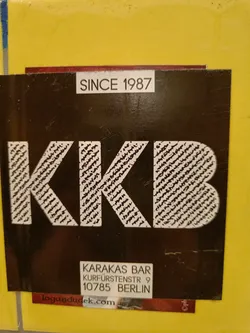 A sticker for the Karakas Bar in Berlin. The sticker is black with white text. The text says "KKB" in large letters, and below it says "Karakas Bar, Kurfürstenstr 9, 10785 Berlin". There is also a website address: logandudek.com and the year 1987.