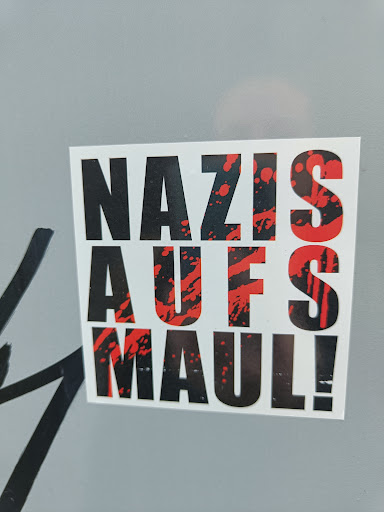 Street sticker A sticker with the text "Nazis Auf's Maul!" in black and red. The text is stylized with red splatters behind it. The sticker is square and printed on white paper.