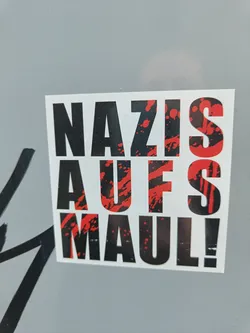 Street sticker A sticker with the text "Nazis Auf's Maul!" in black and red. The text is stylized with red splatters behind it. The sticker is square and printed on white paper.