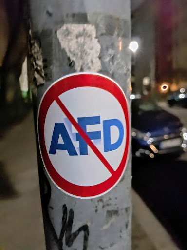 A sticker with a red circle and a diagonal red line crossing out the letters AFD in blue. The sticker is affixed to a gray pole.