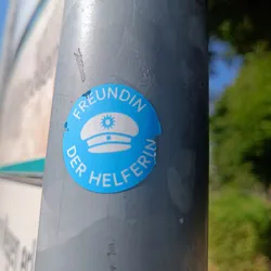 A light blue round sticker with a white police cap in the center and the text "Freundin der Helferin" (Friend of the Helpers) around it.  The sticker is affixed to a gray pole outdoors.