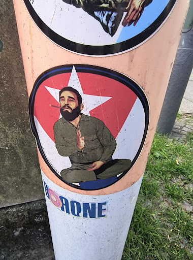 The sticker is round and depicts a man with a beard, resembling Fidel Castro, in a meditative pose with a cigar in his mouth. He is set against a red star on a white and red circular background. The sticker is on a pole, along with other stickers.