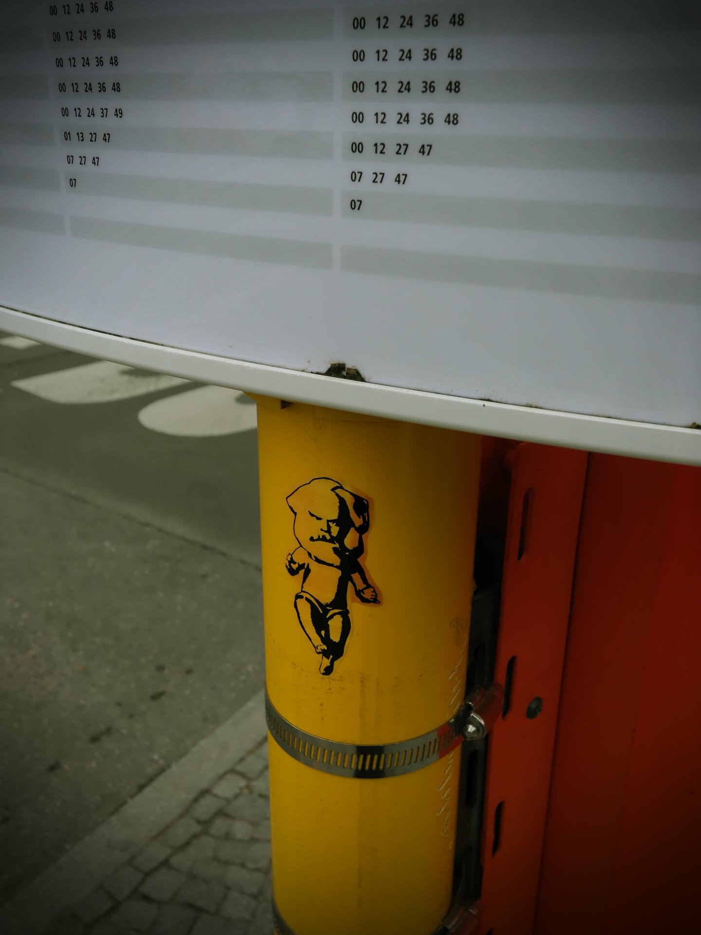 The sticker features a black silhouette of a baby with a prominent, bushy beard and mustache, strongly reminiscent of Karl Marx. The baby is depicted in a dynamic, walking or dancing pose, wearing what appears to be a diaper or briefs. Its facial expression is stern or angry, with furrowed brows. The sticker is applied to a bright yellow cylindrical pole, making the black outline stand out sharply against the vibrant background.