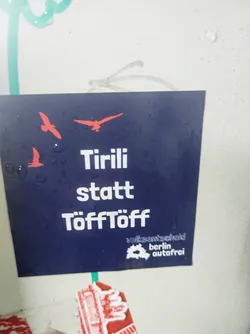 Street sticker Dark blue sticker with three red birds and the text "Tirili statt Töfftöff" (Chirping instead of car noises) in white.  A small logo at the bottom shows a map of Berlin with the text "Volksentscheid Berlin autofrei" (Citizens' initiative Berlin car-free).