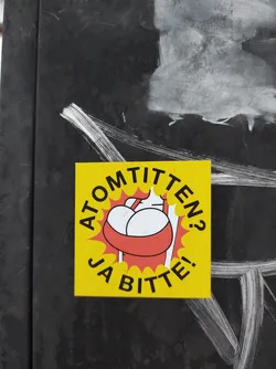 Street sticker Yellow sticker with a cartoon illustration of a person carrying two large breasts in a red basket. The text on the sticker says "Atomtitten? Ja bitte!" which translates from German to "Atomic boobs? Yes please!"