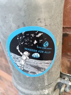 Street sticker A round sticker featuring an illustration of an astronaut on the moon holding up water droplets. The text on the sticker says "VIVA CON AGUA" and "WASSER FÜR ALLE!" which translates to "Water for all!" in German. The sticker has a light blue border and a black background with a small globe image present. 