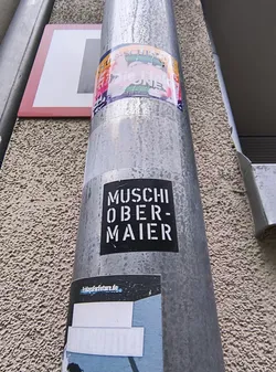 Street sticker The sticker is rectangular with slightly rounded corners, featuring a solid black background. White, stencil-style uppercase letters are prominently displayed, spelling 'MUSCHI' on the top line and 'OBER-MAIER' on the bottom line. The text is centered within the sticker. The sticker shows minor signs of wear, including slight creases and some dirt, indicating its exposure to the elements on the metallic pole.