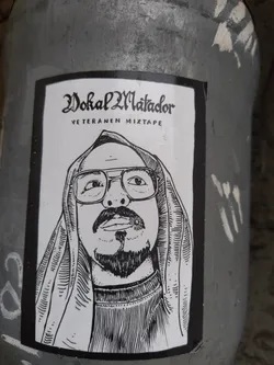 Street sticker The sticker features a black and white illustration of a man's face, centered within a black border. The man is wearing glasses and has a goatee and mustache. He is also depicted wearing a hooded garment, with the hood pulled up over his head. The style of the illustration is detailed, with fine lines used to create shading and texture, particularly on the clothing and facial hair. Above the portrait, in a stylized gothic font, is the text 'Vokal Matador', and below that, in a simpler font, is 'VETERANEN MIXTAPE'.