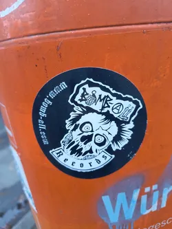 Street sticker The sticker is circular and predominantly black and white. It features a stylized skull with wide, staring eyes and spiky hair. Above the skull, the text 'BOMB' is prominently displayed, with a bomb graphic integrated into the lettering. To the right of 'BOMB' is the anarchy symbol (a circle with an 'A' inside) and an image of two crossed bones or axes. The sticker also has text curving around the bottom edge, reading 'Records' within a banner. Along the top edge, in a smaller font, is the URL 'www.bomb-a-ll.com'. The overall aesthetic is gritty and punk-inspired.