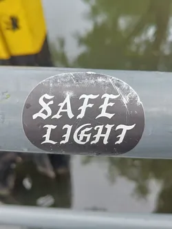 A round black and white sticker with the words "SAFE LIGHT" written in a stylized font.  The sticker is affixed to a metal railing outdoors.