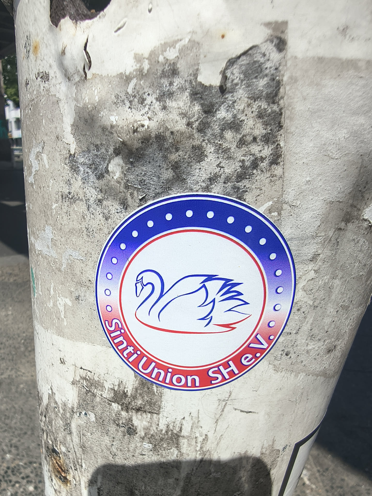 A round sticker on a textured pole. It depicts a stylized blue and red swan inside a white circle. The outer ring has a blue-to-red gradient, white dots, and the text 'Sinti Union SH e.V.'.