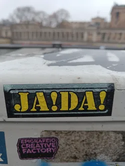 Street sticker A yellow and black sticker with the text "JA!DA!" in bold letters. The sticker is affixed to a white surface and appears to be outdoors. There are water droplets visible on the sticker's surface.  Below it is another sticker that reads "EPICRAFEIO CREATIVE FACTORY".