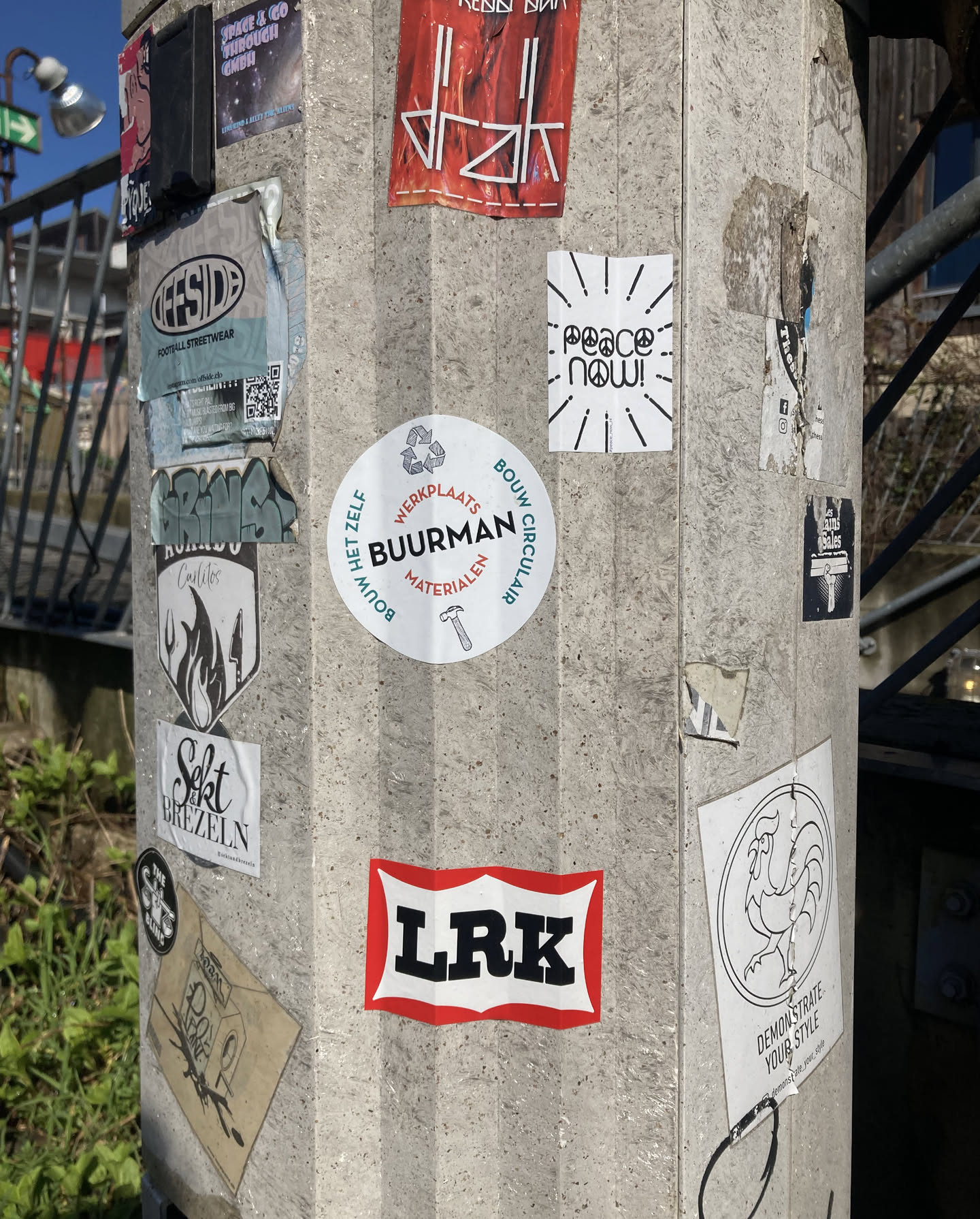 The central focus is a collection of stickers adhered to a concrete pillar. One prominent circular sticker is white with blue and red text and a small hammer icon at the bottom. It reads 'BUURMAN MATERIALEN' with 'WERKPLAATS' and 'BOUW CIRCULAIR' arching above and below. To its upper right is a rectangular sticker with black and white lines radiating outwards, and the text 'peace now!' in a playful font, with peace symbols for the 'o's. Below the 'peace now!' sticker and to the left of the 'BUURMAN' sticker is a black and white shield-shaped sticker with flames and a chef's hat, with 'Carlitos' written above the flames. Below that, a smaller white sticker reads 'Scht BREZELN' in stylized text. To the right of the 'BUURMAN' sticker, a larger sticker features a stylized rooster in a circle with text below. Another sticker to the left of the 'BUURMAN' sticker, partially obscured, seems to have graffiti-style writing. Further up, a red sticker with white text is visible. A blue and white sticker for 'OFFSIDE FOOTBALL STREETWEAR' is also present, containing a QR code. Another sticker to the right of the 'peace now!' sticker has a black background with white text and imagery, and another social media icon is visible below it. At the bottom right, a sticker with a red border and white text reads 'LRK' in bold black letters.