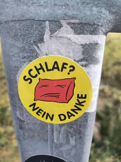 Street sticker A round yellow sticker with a red cartoon pillow with x eyes and a tongue. The text around it says "Schlaf? Nein Danke" which translates from German to "Sleep? No thanks!"