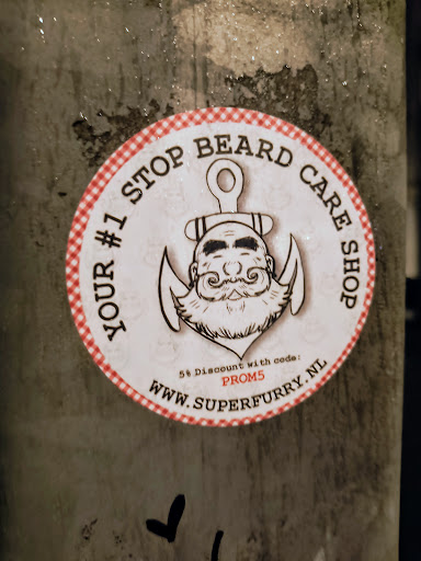 Street sticker A round sticker with a red and white checkered border features an illustration of a bearded man with an anchor above his head, advertising "YOUR #1 STOP BEARD CARE SHOP". A promotional offer is mentioned at the bottom: "5% Discount with code: PROM5", along with the website address "WWW.SUPERFURRY.NL".