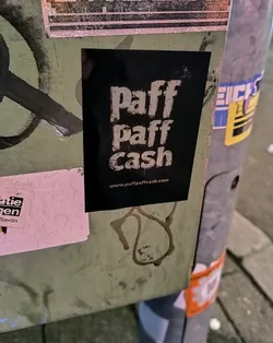 Street sticker The sticker is black with white text. It features the words 'Paff Paff Cash' stacked on top of each other. Below the words is the website address 'www.paffpaffcash.com'. The sticker is adhered to a pole with other stickers and marks.
