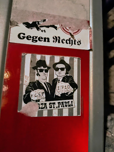 A sticker depicting two men in suits, reminiscent of the Blues Brothers, with knuckles showing FCSP and 1910. The background features vertical stripes and the text "Forza St. Pauli".  There is also another sticker above it with the text "Gegen Rechts" visible.